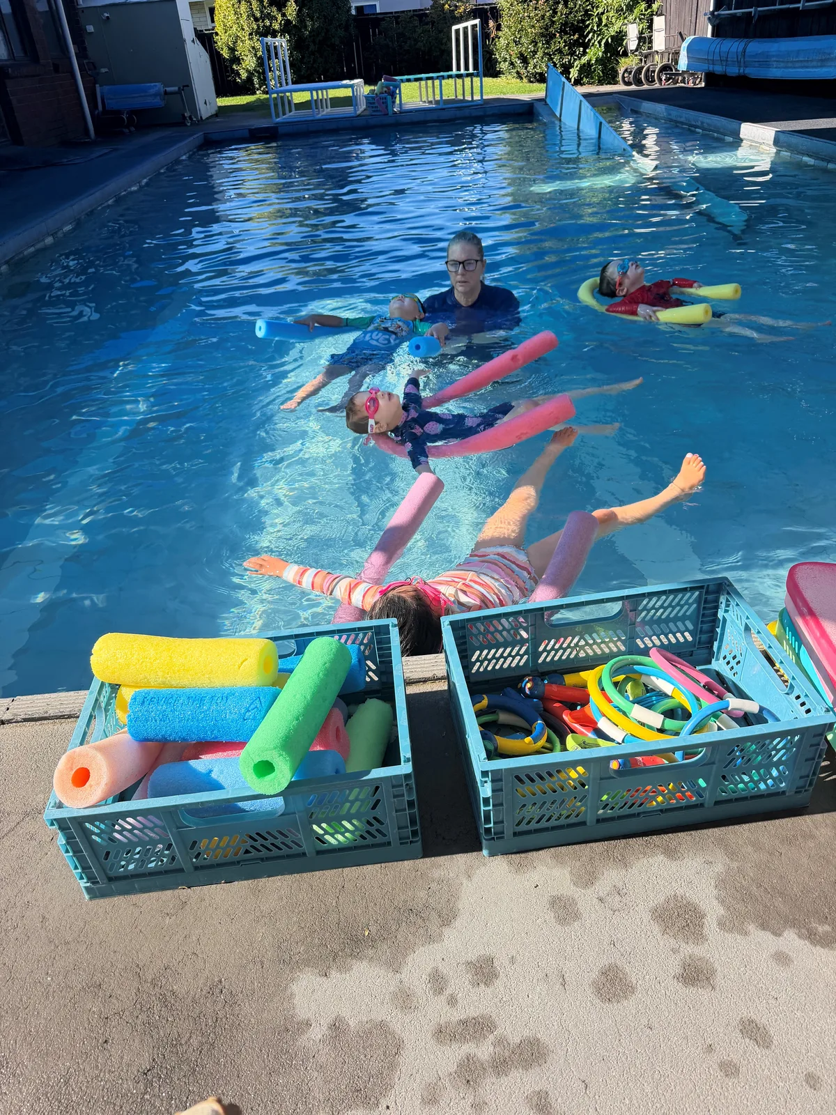 Children swimming with pool noodles and a teacher at a swimming session