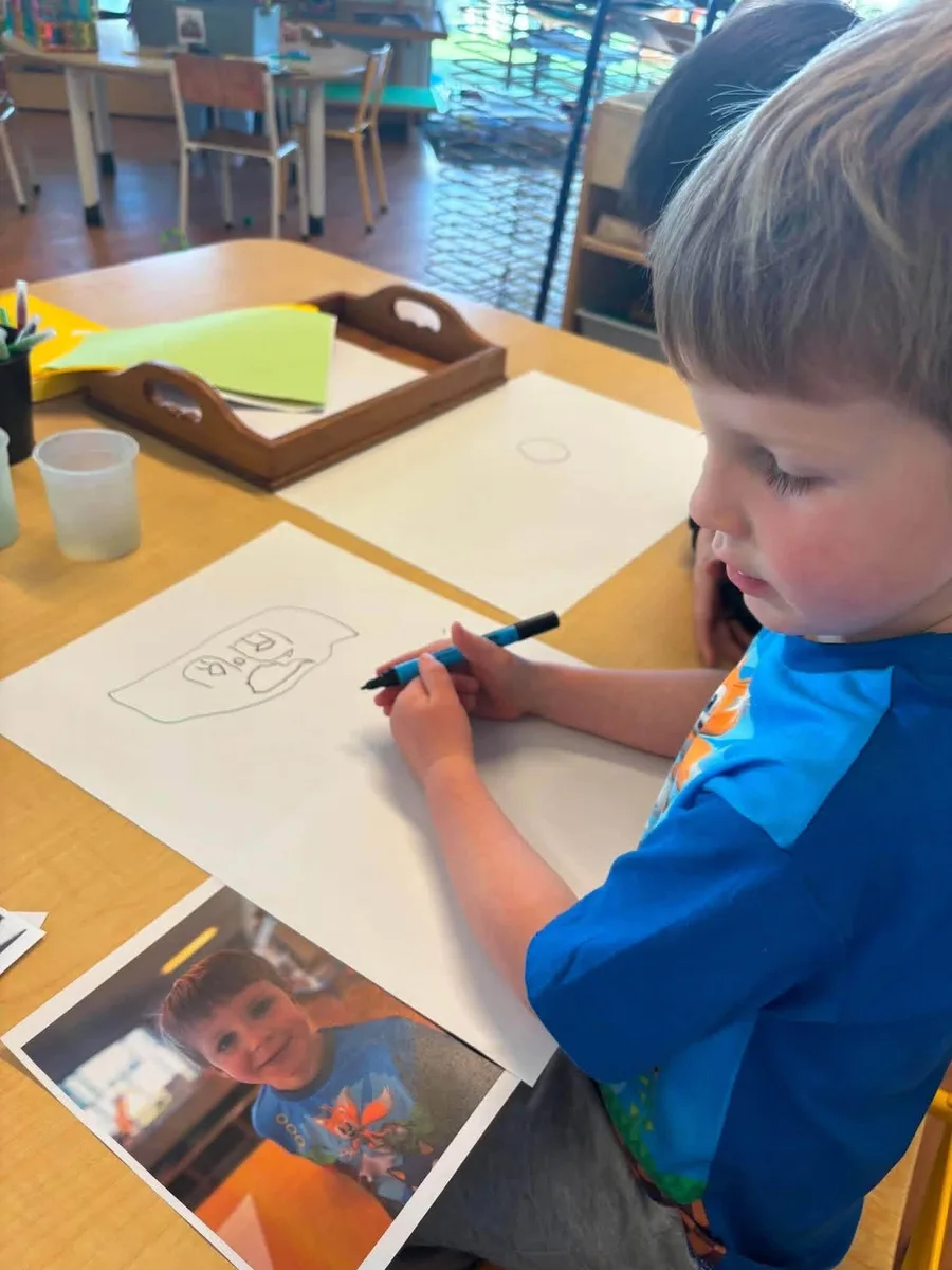 A preschooler drawing a self portrait at a table with felt pens